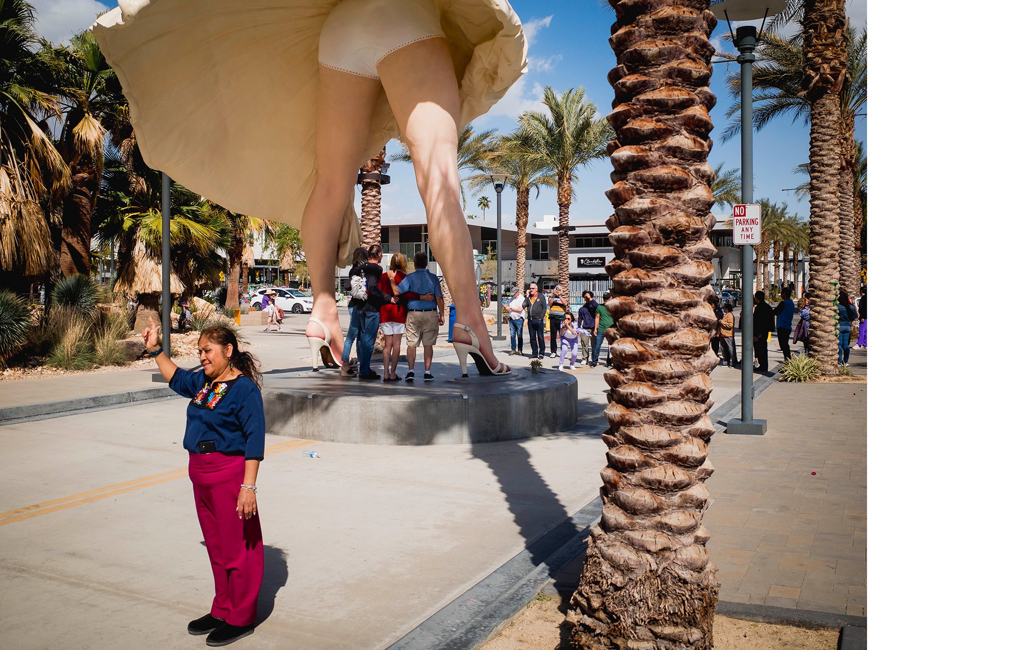 A woman in a blue top and pink pants stands in front of a large statue of a woman in high heels. The statue's legs and skirt are visible. The statue is placed on a circular platform outdoors, surrounded by palm trees and a group of people.