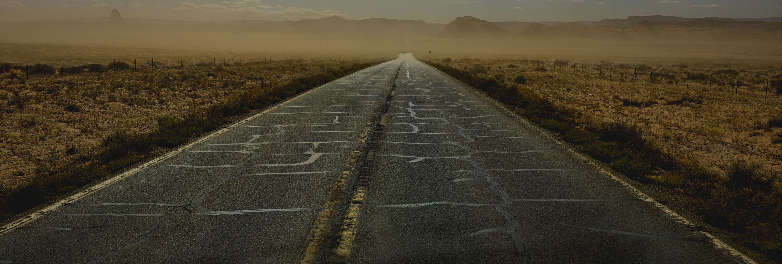 A long, straight road running through a desert landscape with mountains in the distance and a fence on either side, under a hazy sky.