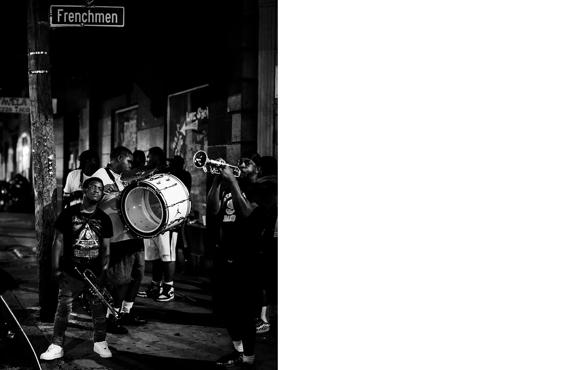 A group of young musicians playing instruments on a city street at night. One youth is holding a trumpet, one holds a drum, and others are gathered around. Street sign reads 'Frenchmen' and there are posters on the wall behind them.