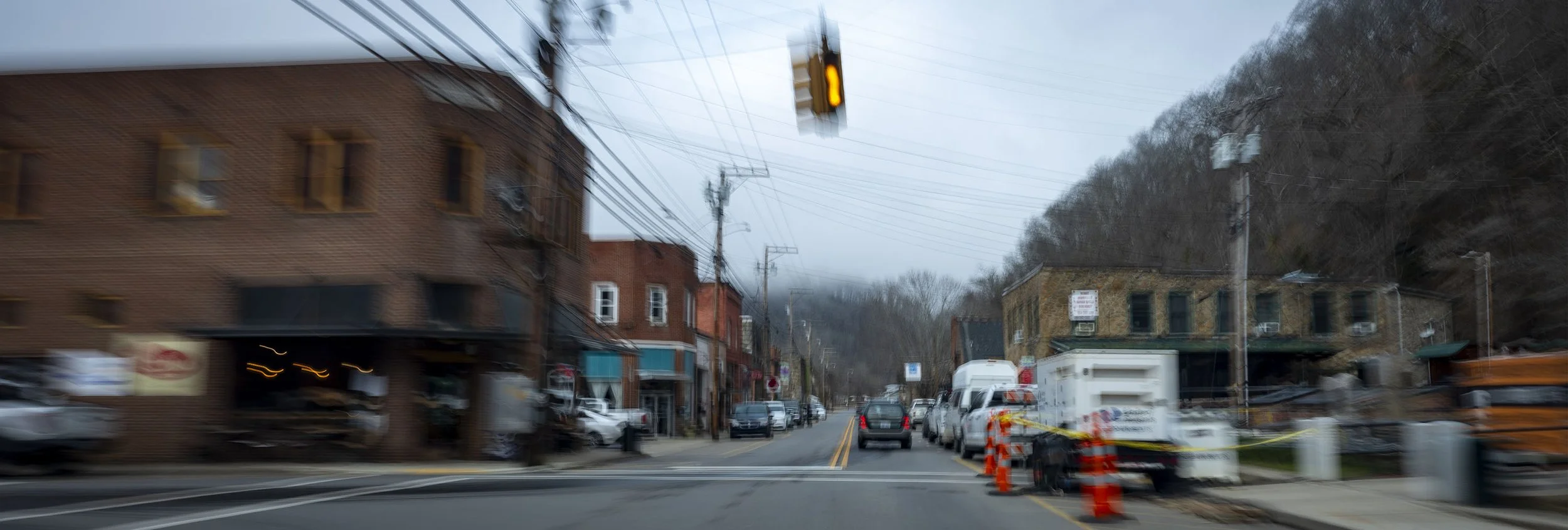 Blurred image of a small town street during daytime with traffic lights, parked cars, and buildings on either side. Construction cones and equipment are visible on the right side.