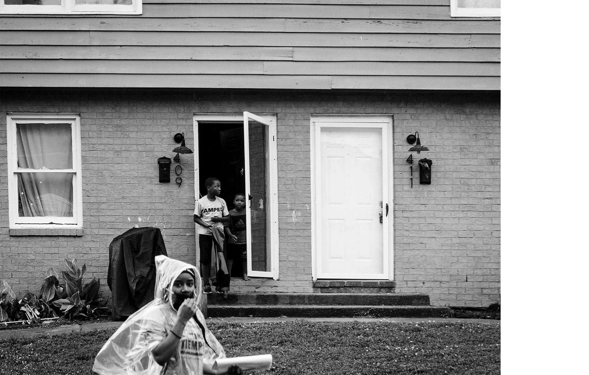 Black and white photo of a brick house with two children standing on the steps near the open front door. One child is wearing a t-shirt with the word "CAMPE" visible. A woman in a raincoat and face mask is sitting on the grass in the foreground. Wind