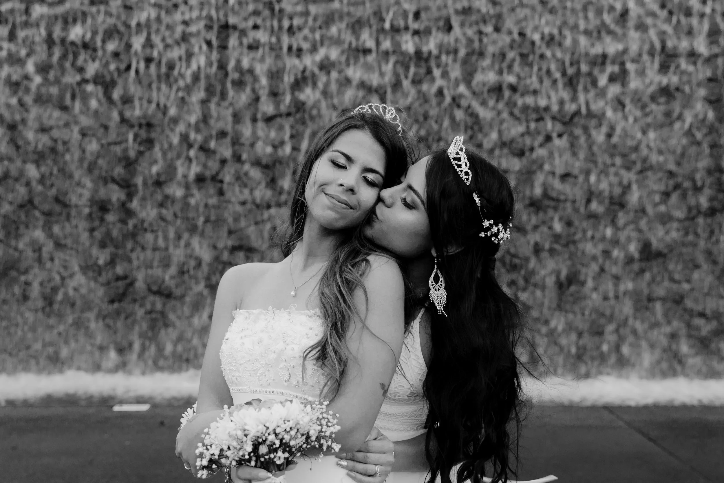 Two women dressed in wedding attire sharing a kiss outdoors, one holding a bouquet of flowers, with a stone wall in the background.