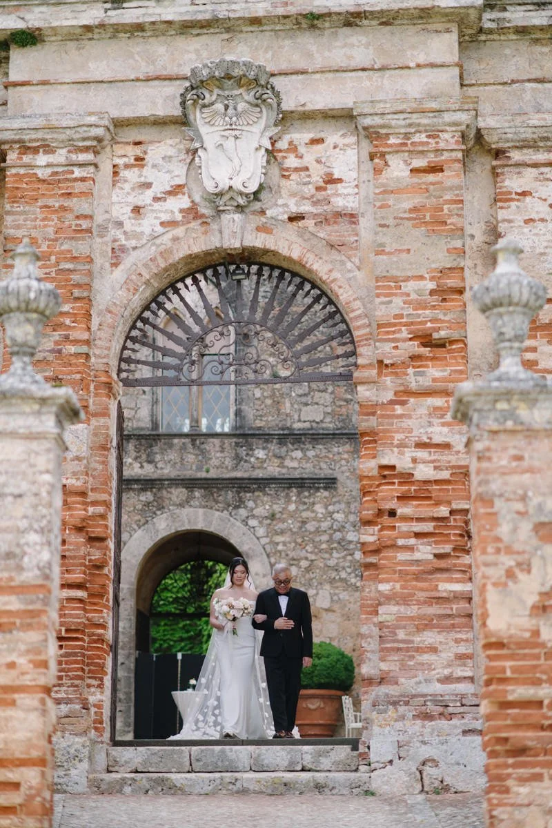 Bride walking with her father through the main entrance of Castello di Celsa before the wedding ceremony.