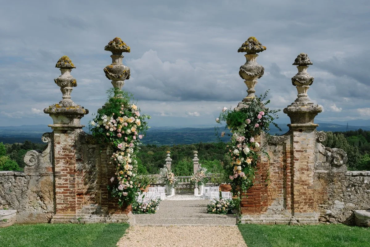 Ceremony area at Castello di Celsa seen from a distance, framed by stone pillars and rolling Tuscan landscape.
