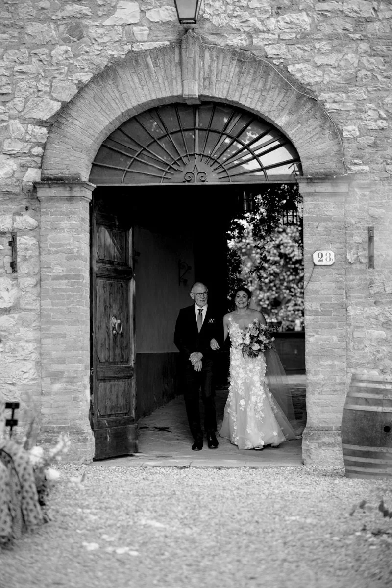 Black and white photo of the bride walking with her father through the castle doorway before the wedding ceremony at Castello di Bossi in Tuscany.