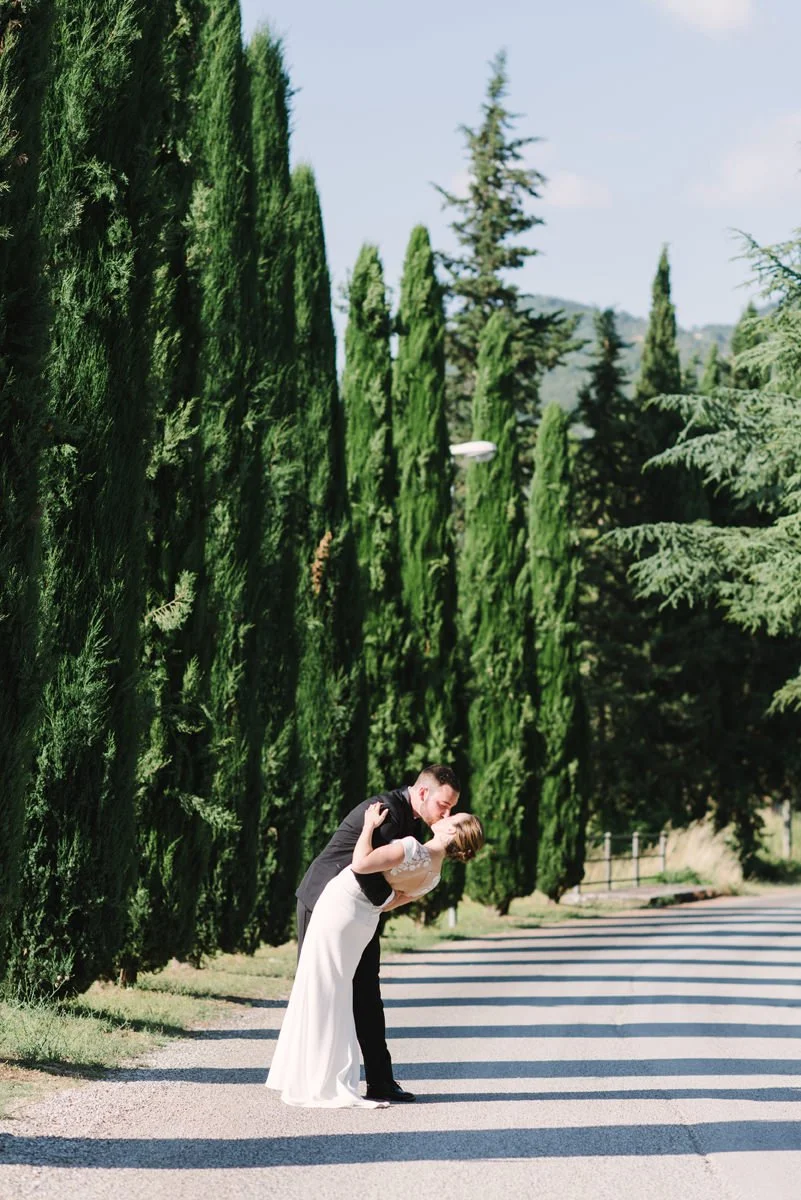 Wedding couple kissing on the road leading to Castello di Spaltenna, framed by tall Tuscan trees.