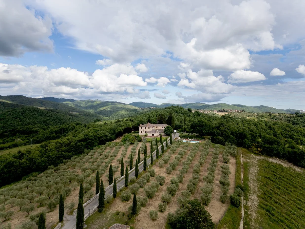 Drone view of Castello di Meleto showing house buildings, surrounding vineyards, and cypress-lined roads in the Chianti countryside.