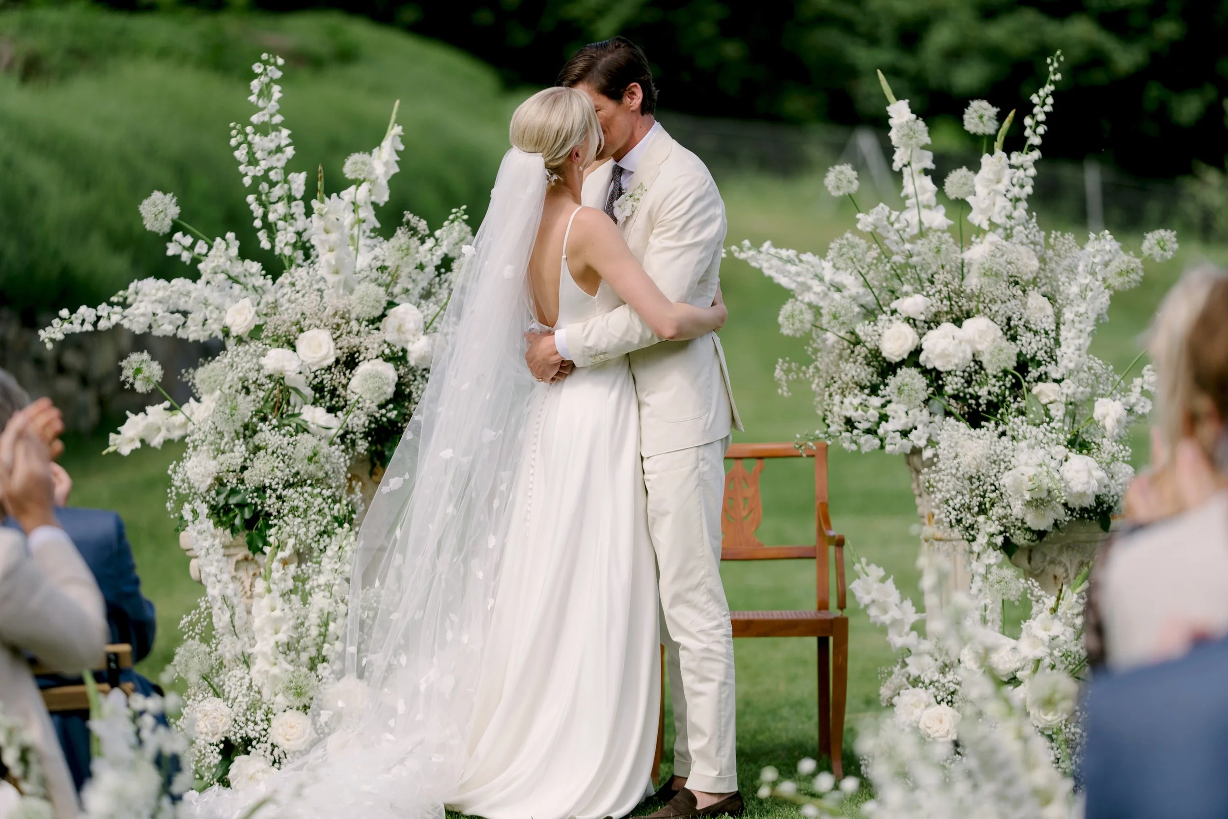 Bride and groom embracing beside an arch of white flowers and gypsophila during the ceremony at Spao Borgo San Pietro, Umbria