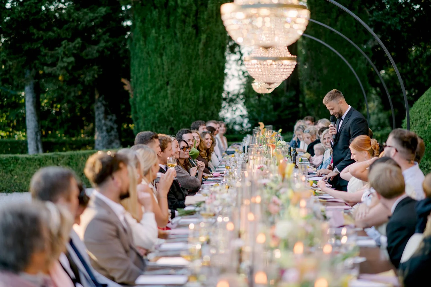 Wedding guests seated and groom is giving speech at the long table with chandeliers and candles in front of Villa di Ulignano, October Tuscany dinner by FunkyBird Photography