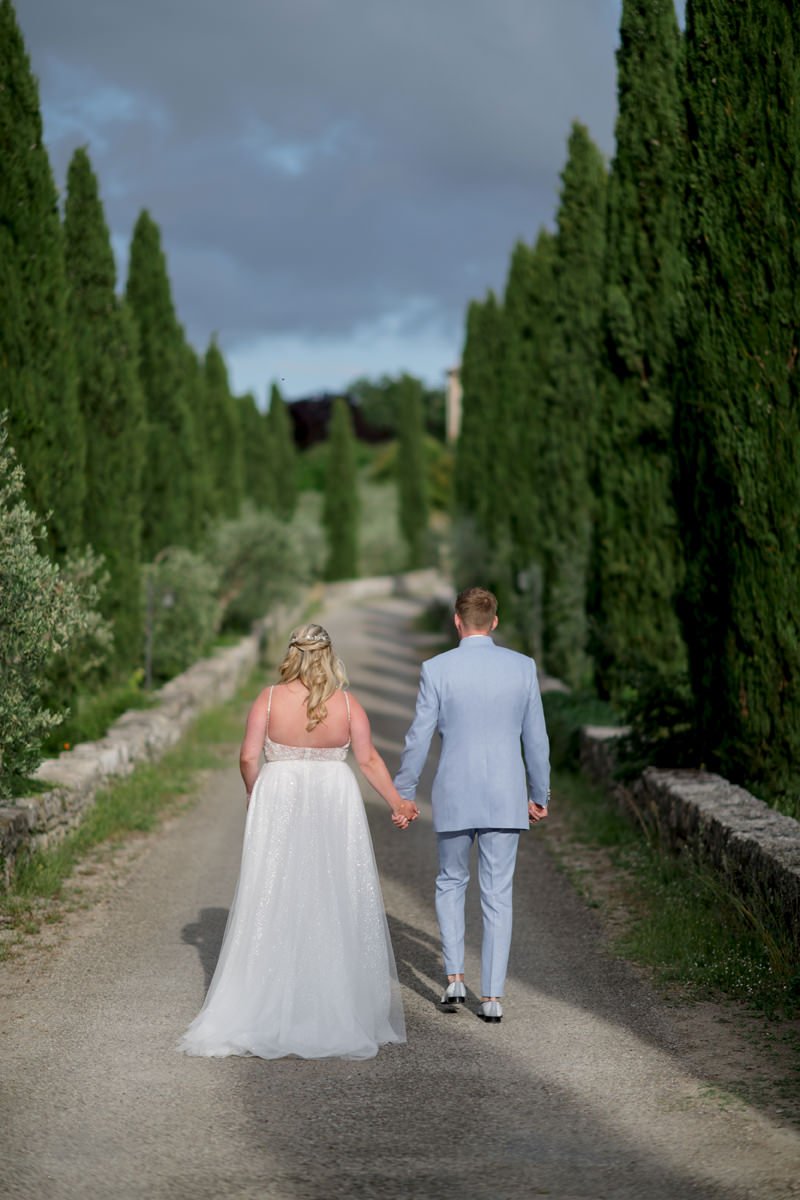Wedding couple walking between tall cypress trees on the grounds of Castello di Meleto in Tuscany.