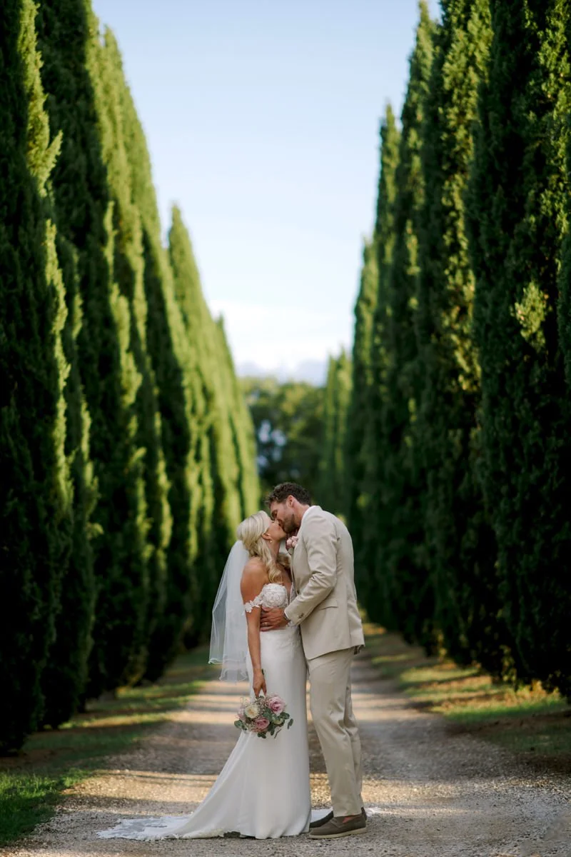 Bride and groom kissing between tall Tuscan cypress trees near Villa Boscarello.