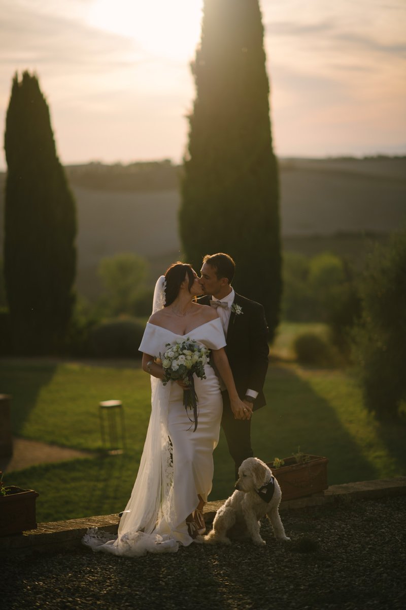 Bride and groom kissing during sunset at Villa Boscarello while a small dog stands beside them in the Tuscan countryside.