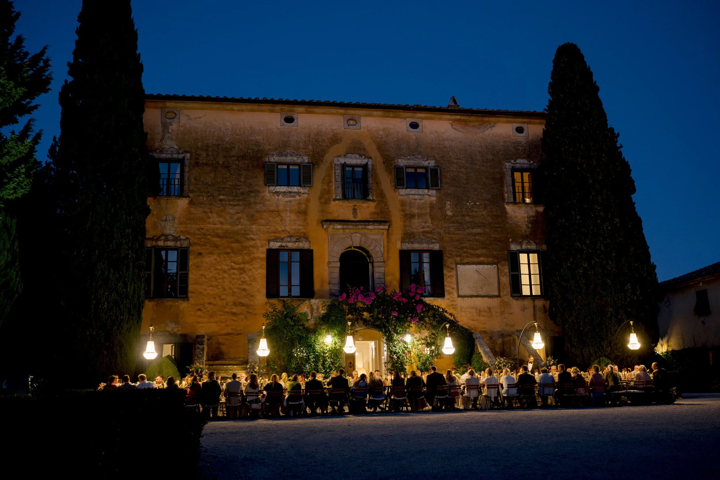 Long-table wedding dinner with six crystal chandeliers in front of Villa di Ulignano at blue hour, October Tuscany wedding photographed by FunkyBird Photography