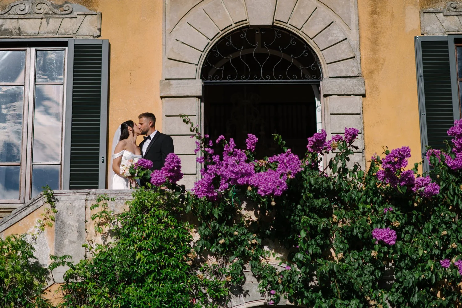 Bride and groom kissing on the front stairs of Villa di Ulignano framed by flowering bougainvillea, by FunkyBird