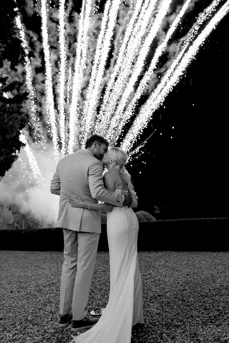 Black and white photo of the couple kissing while fireworks explode in the sky during their wedding celebration at Villa Boscarello.
