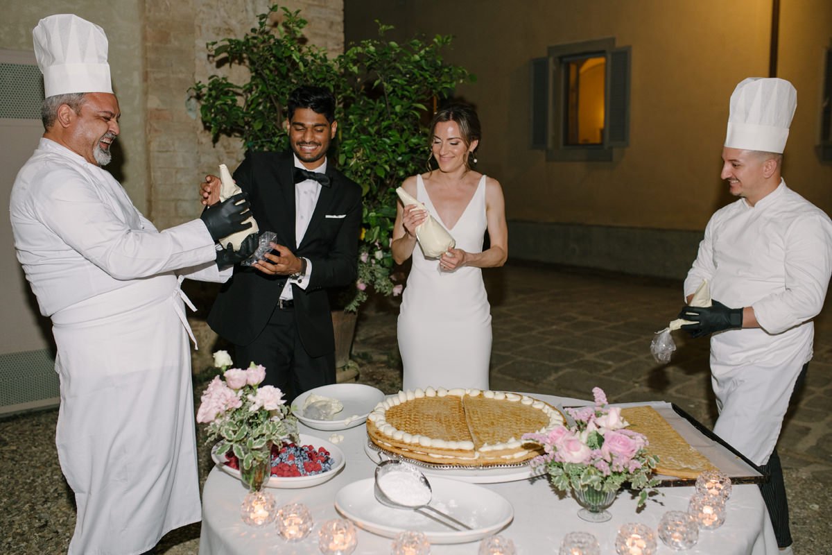 Bride and groom smiling with chefs preparing a millefoglie wedding cake at Tenuta di Monaciano.