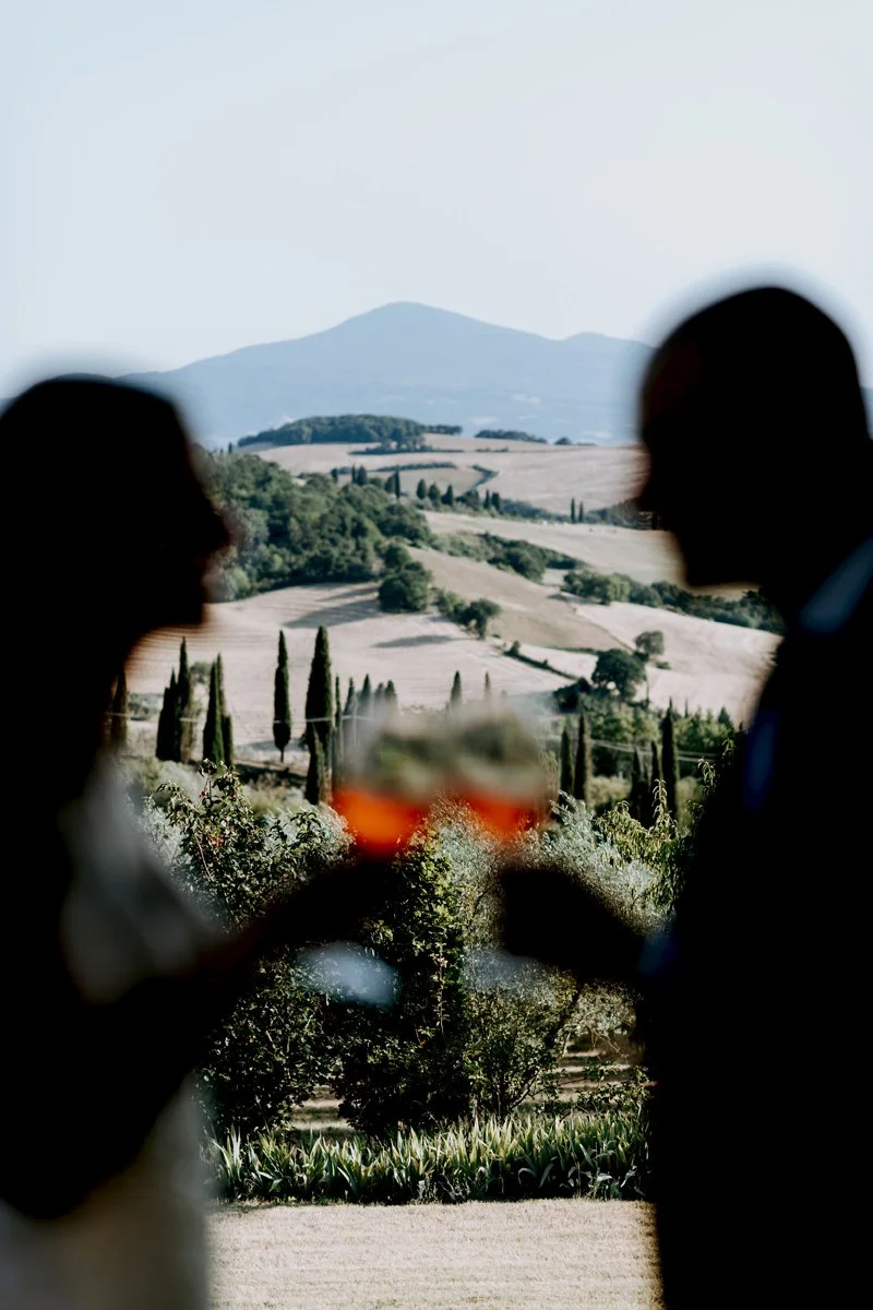 Shadow of a wedding couple cheering with Aperol Spritz during aperitivo at Terre di Nano with Tuscan hills behind.