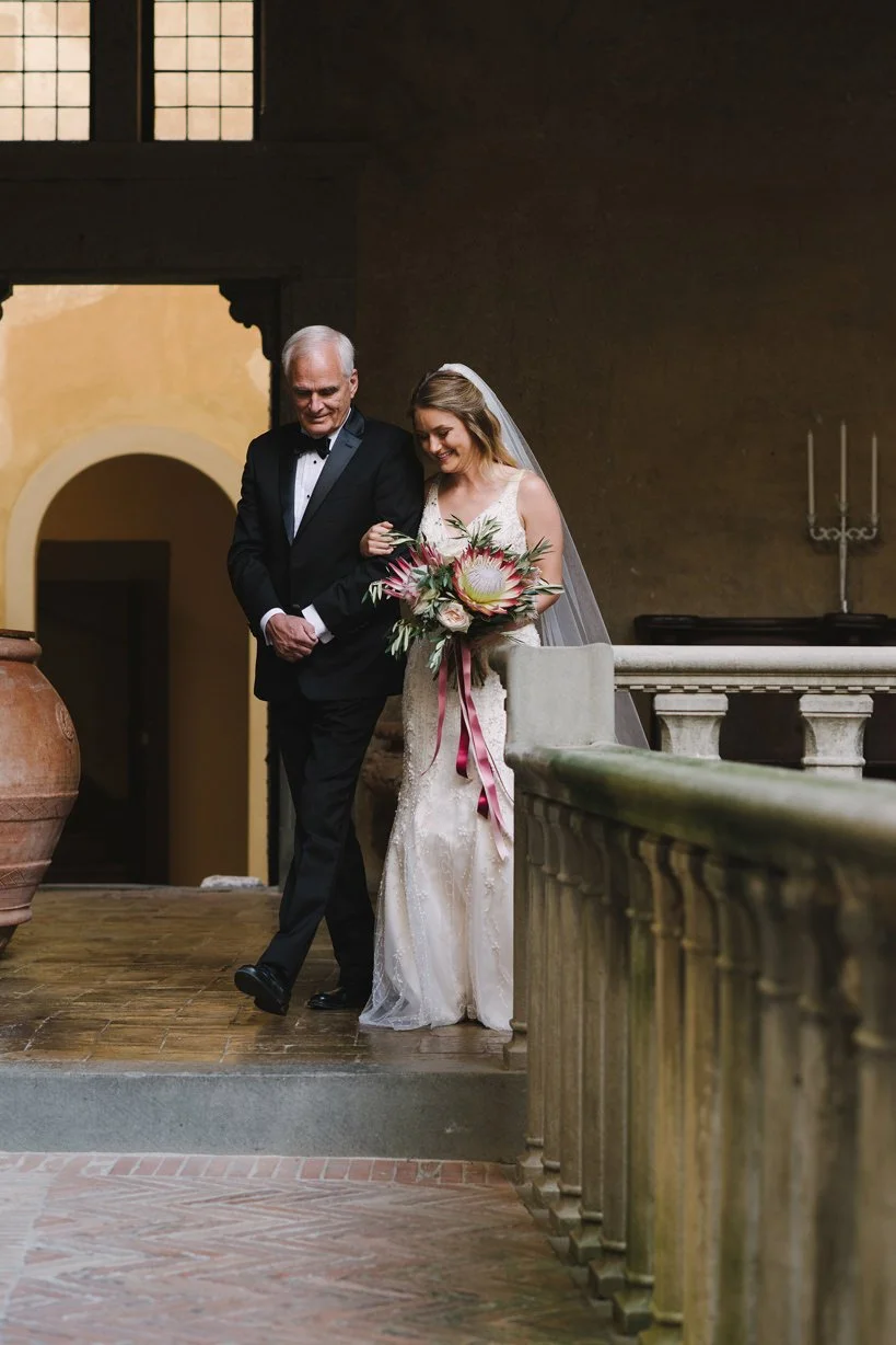 Bride walking with her father toward the ceremony at Castello di Montegufoni, inside the historic courtyard space.