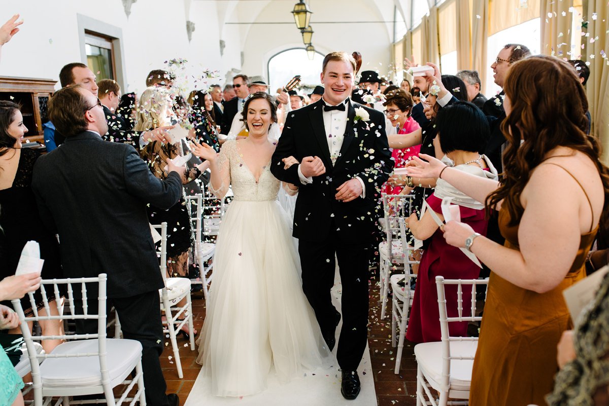 Newly married couple smiling while guests throw petals after the ceremony at Villa Hotel Tolomei.