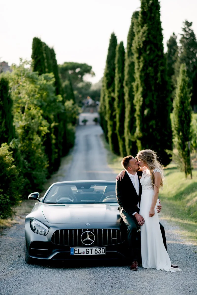 Couple kissing on a modern Mercedes cabrio at Terre di Nano, with tall Tuscan cypress trees in the background.