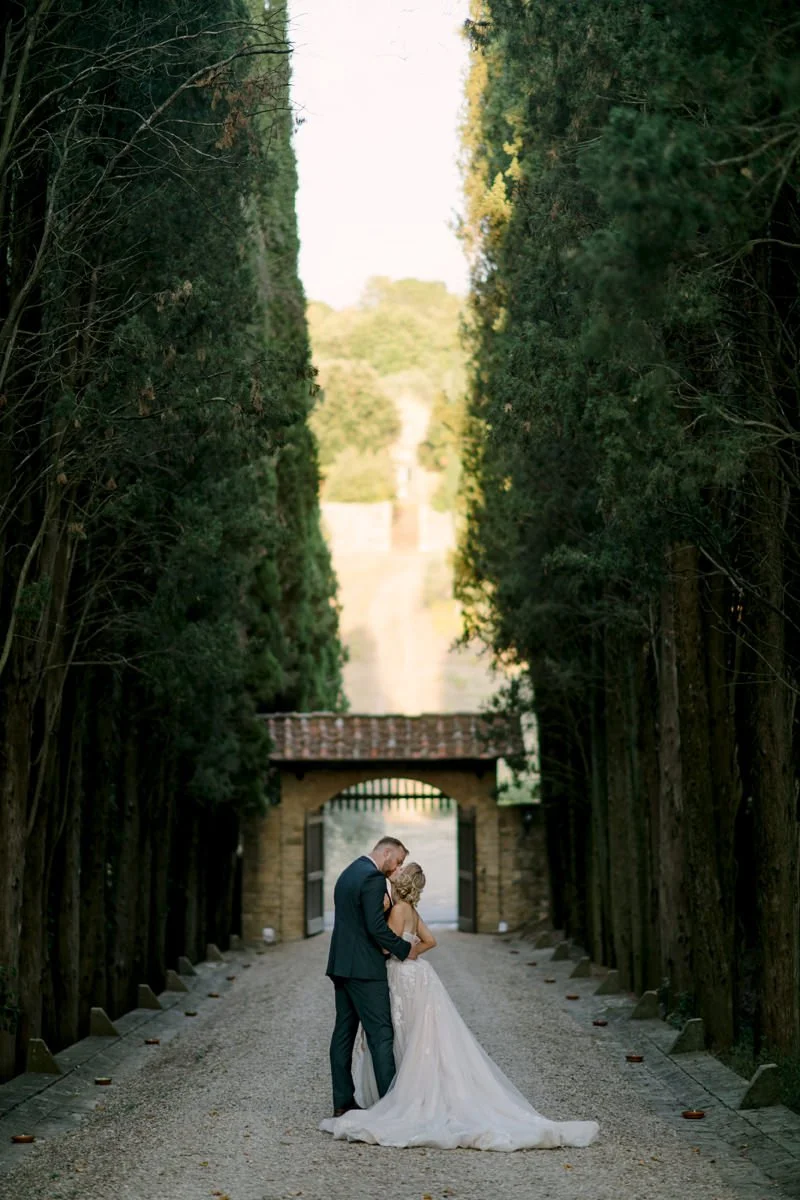 Bride and groom kissing on the road leading to Castello Il Palagio surrounded by tall Tuscan trees.