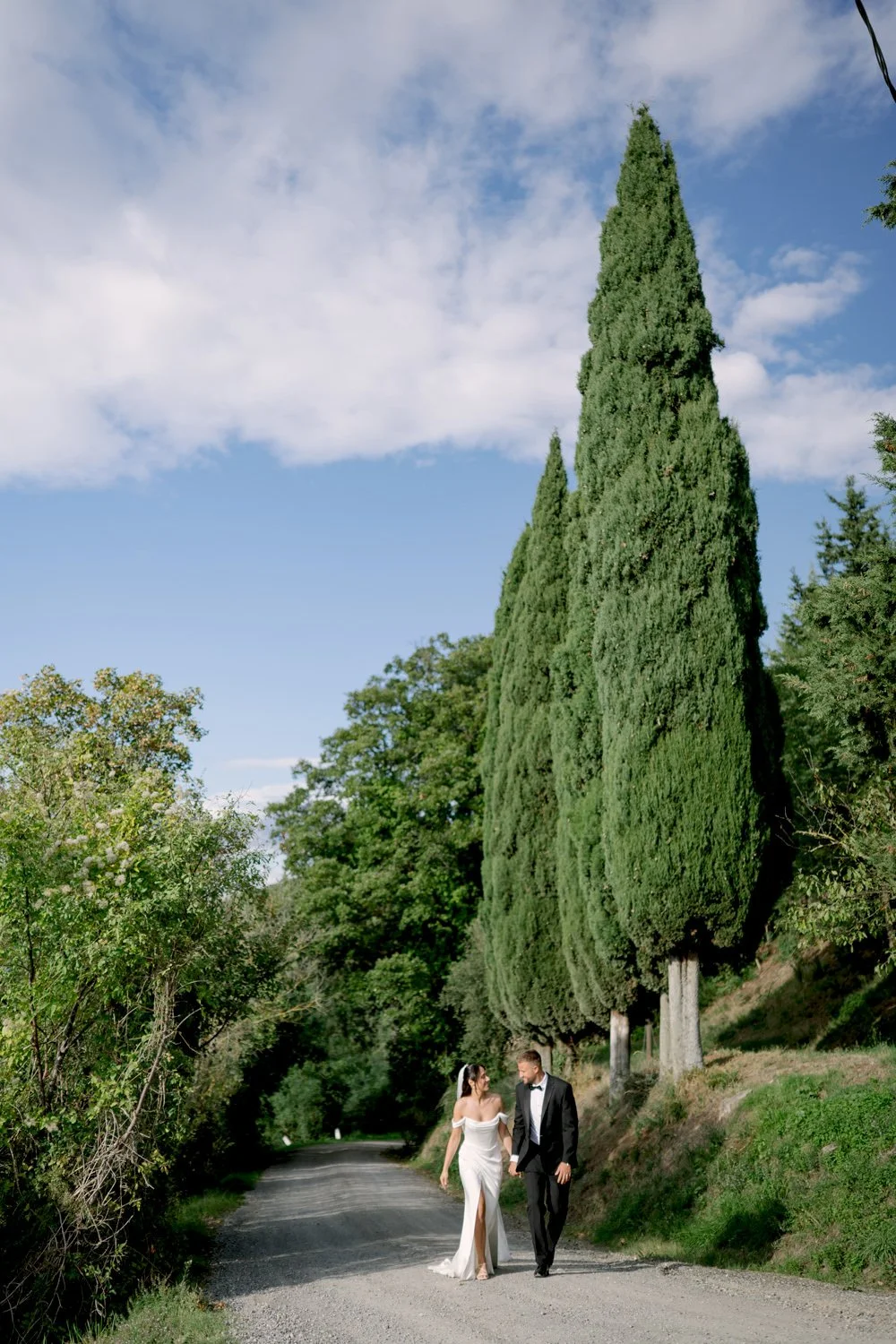 Bride and groom walking past tall Tuscan cypress trees at Villa di Ulignano, October wedding portrait by FunkyBird