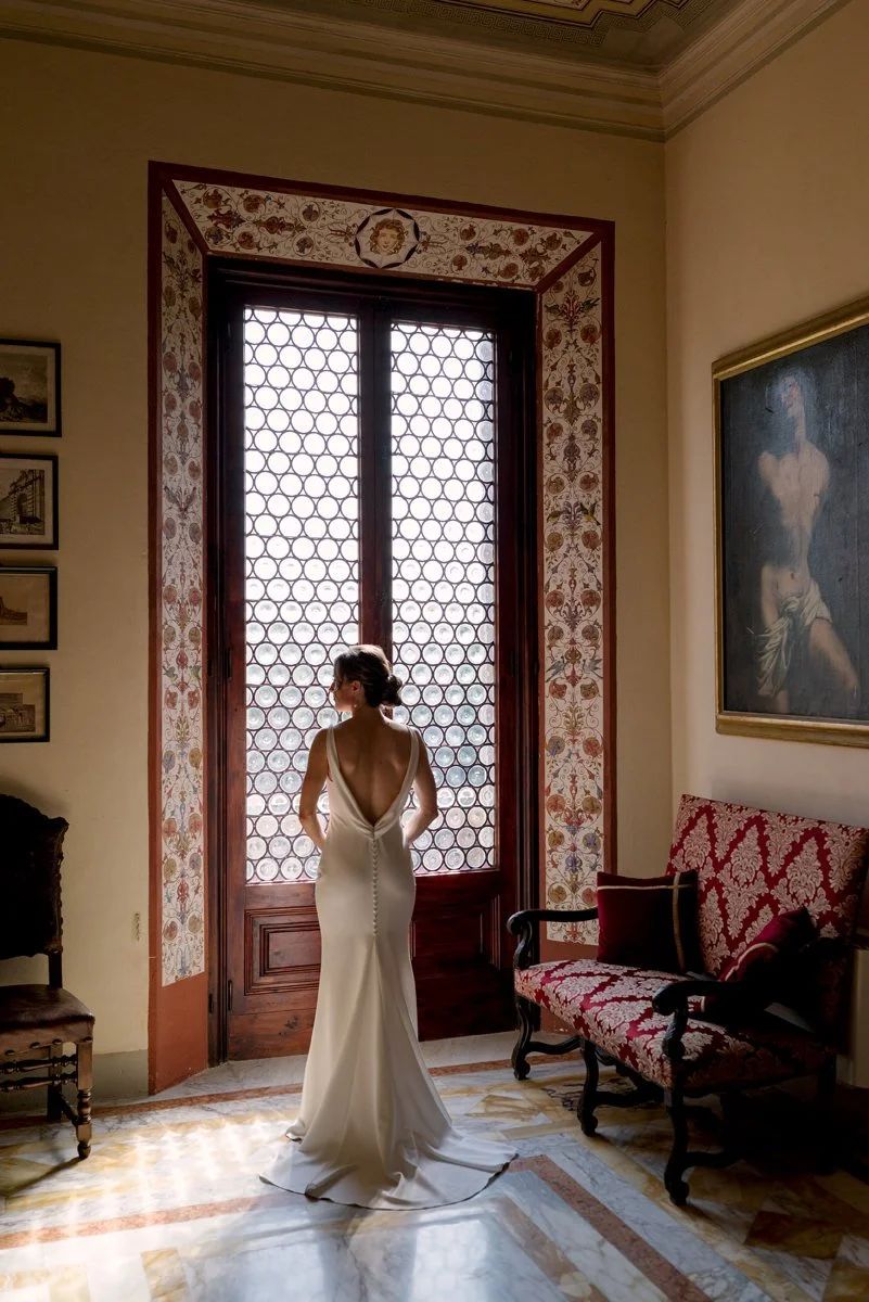 Bride standing with her back to a large window inside Tenuta di Monaciano, soft natural light filling the room.