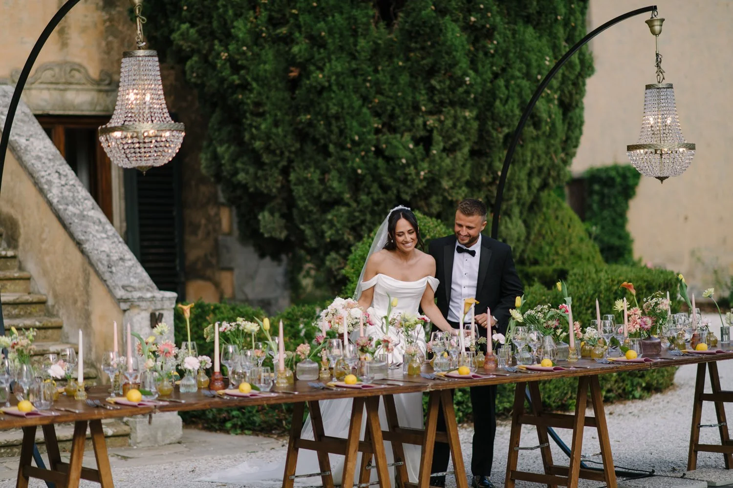 Bride and groom at the long dinner table in front of Villa di Ulignano with chandeliers above, by FunkyBird