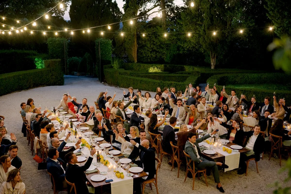 Wedding dinner in front of Villa di Ulignano with multiple long tables as guests cheer and celebrate together.