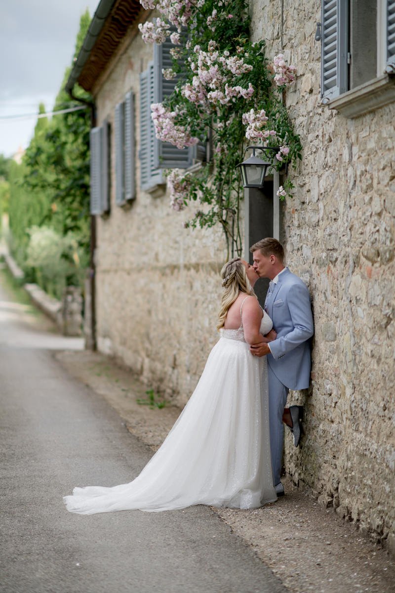 Wedding couple kissing beside a stone house at Castello di Meleto, surrounded by hanging flowers in the Chianti countryside of Tuscany.