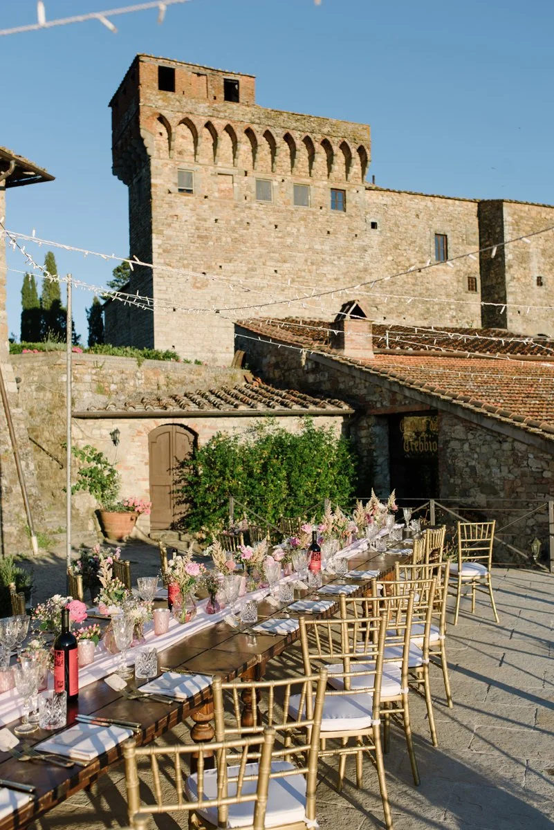 Long wedding dinner table set in front of Castello del Trebbio with historic stone architecture as backdrop.
