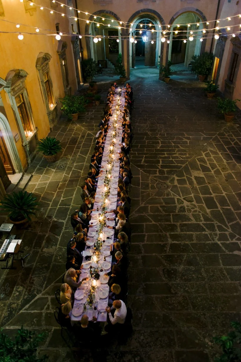 Long wedding dinner table set inside the courtyard of Castello di Montegufoni with candles and floral decor.