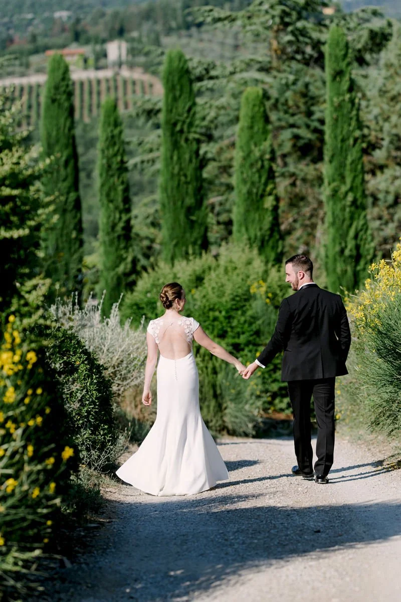 Wedding couple walking across the grounds of Castello di Spaltenna with tall Tuscan trees surrounding them.