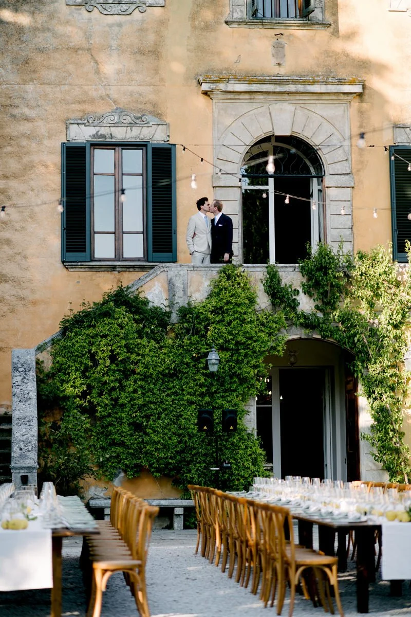 Same sex couple kissing on the stairs at Villa di Ulignano, with wedding dinner tables set in front of the villa.