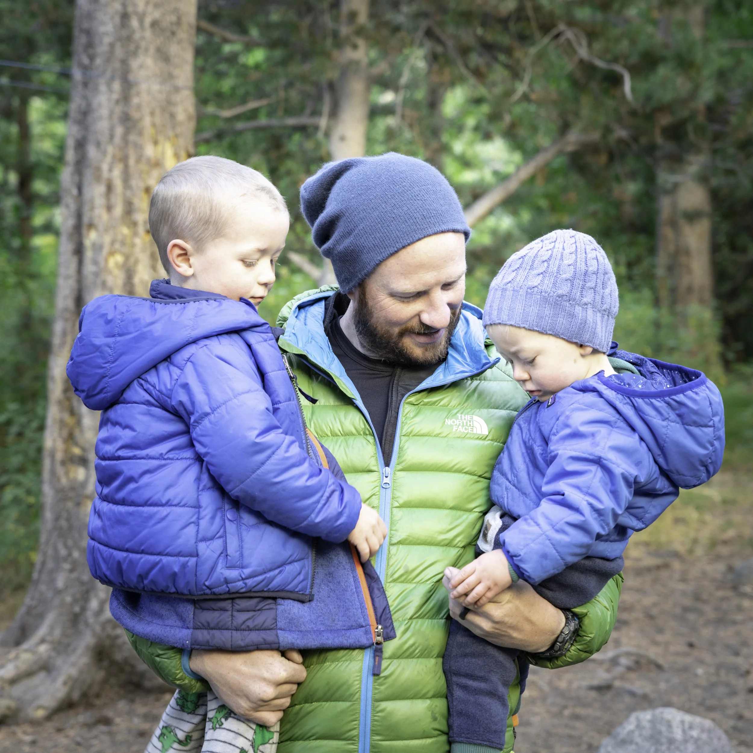 Jason White, owner of Apex Construction & Restoration, holding his two sons while camping in a forest setting in California.