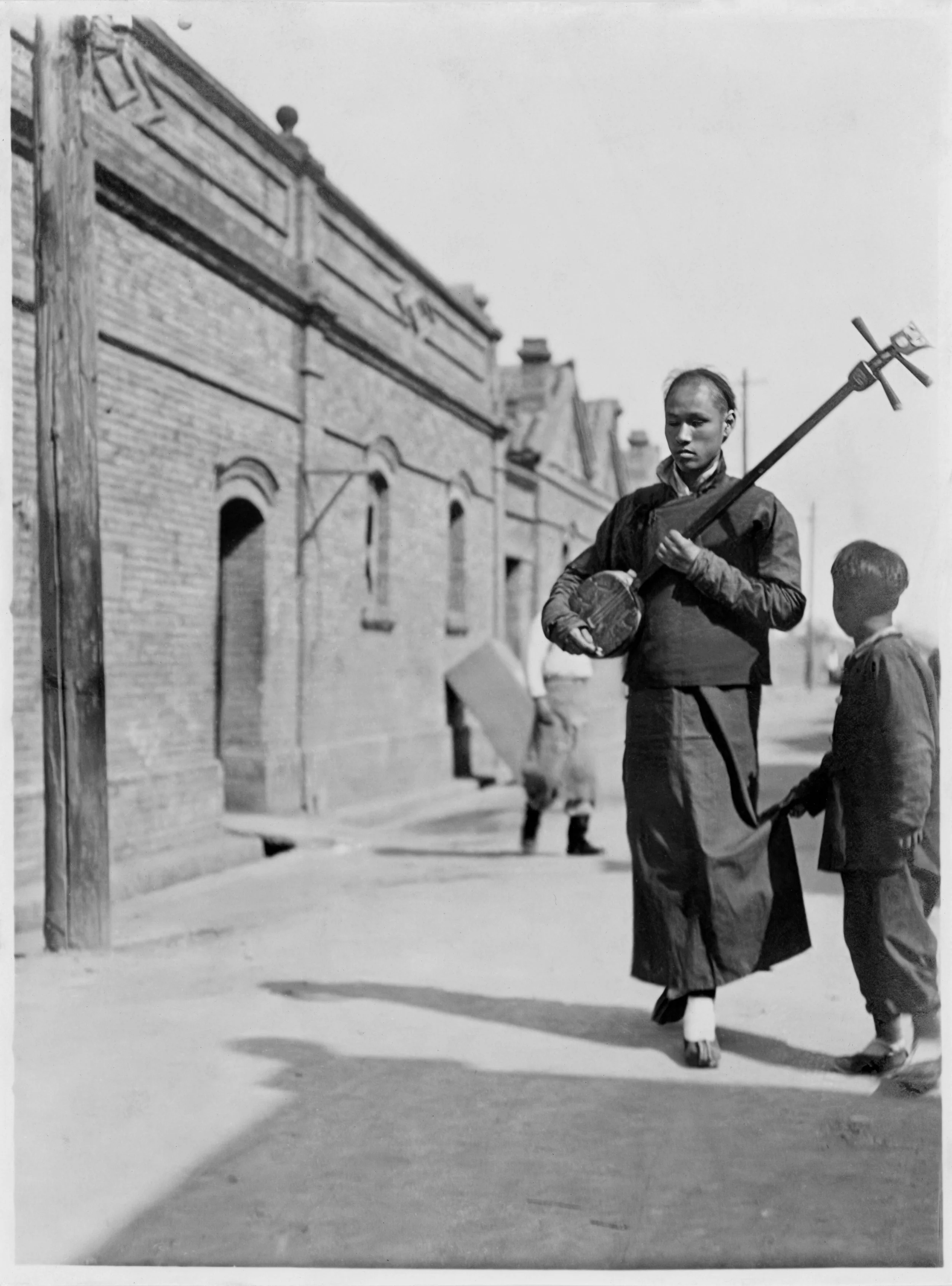 A mistral and his son walk in front of a brick building. The boy looks back at a soldier.