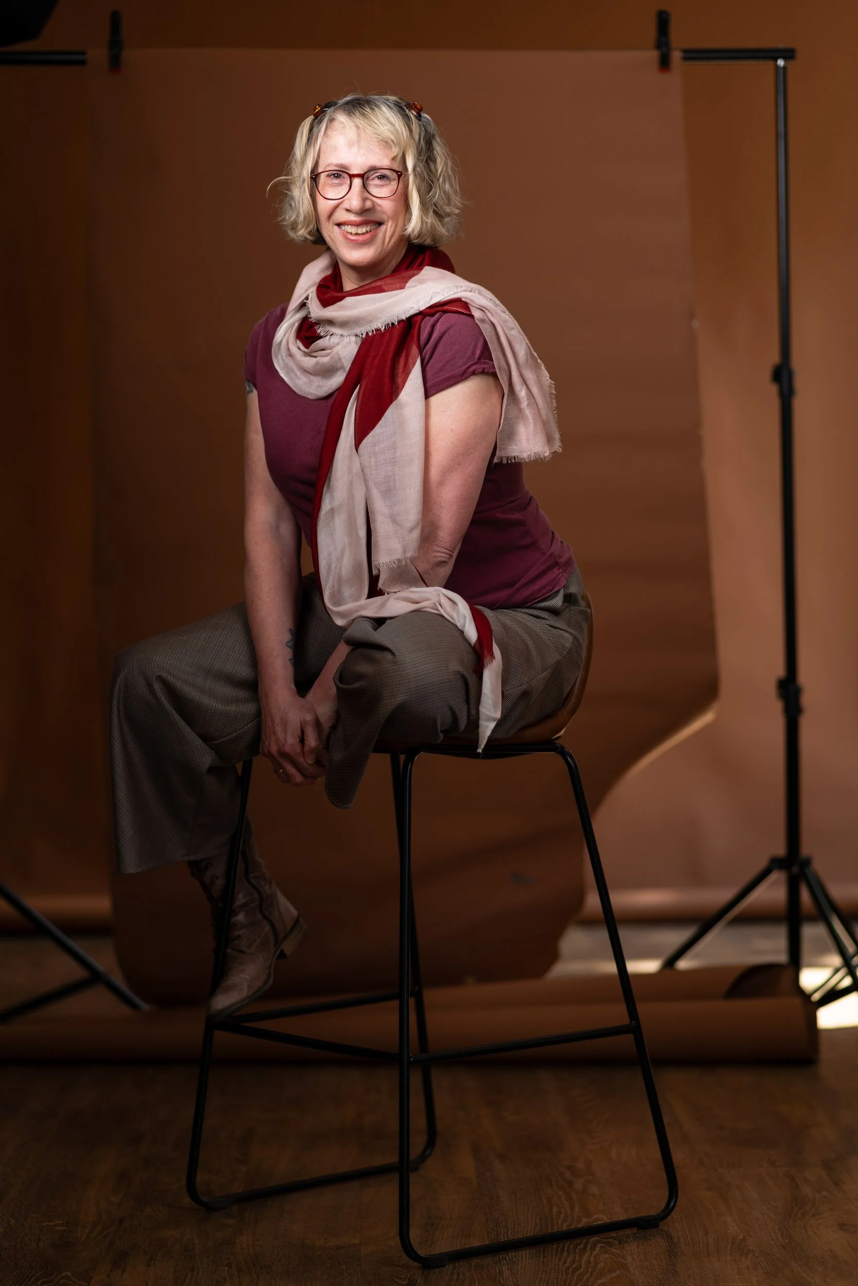Pauline Klemm photographer sits on a stool for a portrait. She is relaxed and smiling. A scarf is casually wrapped around her neck.