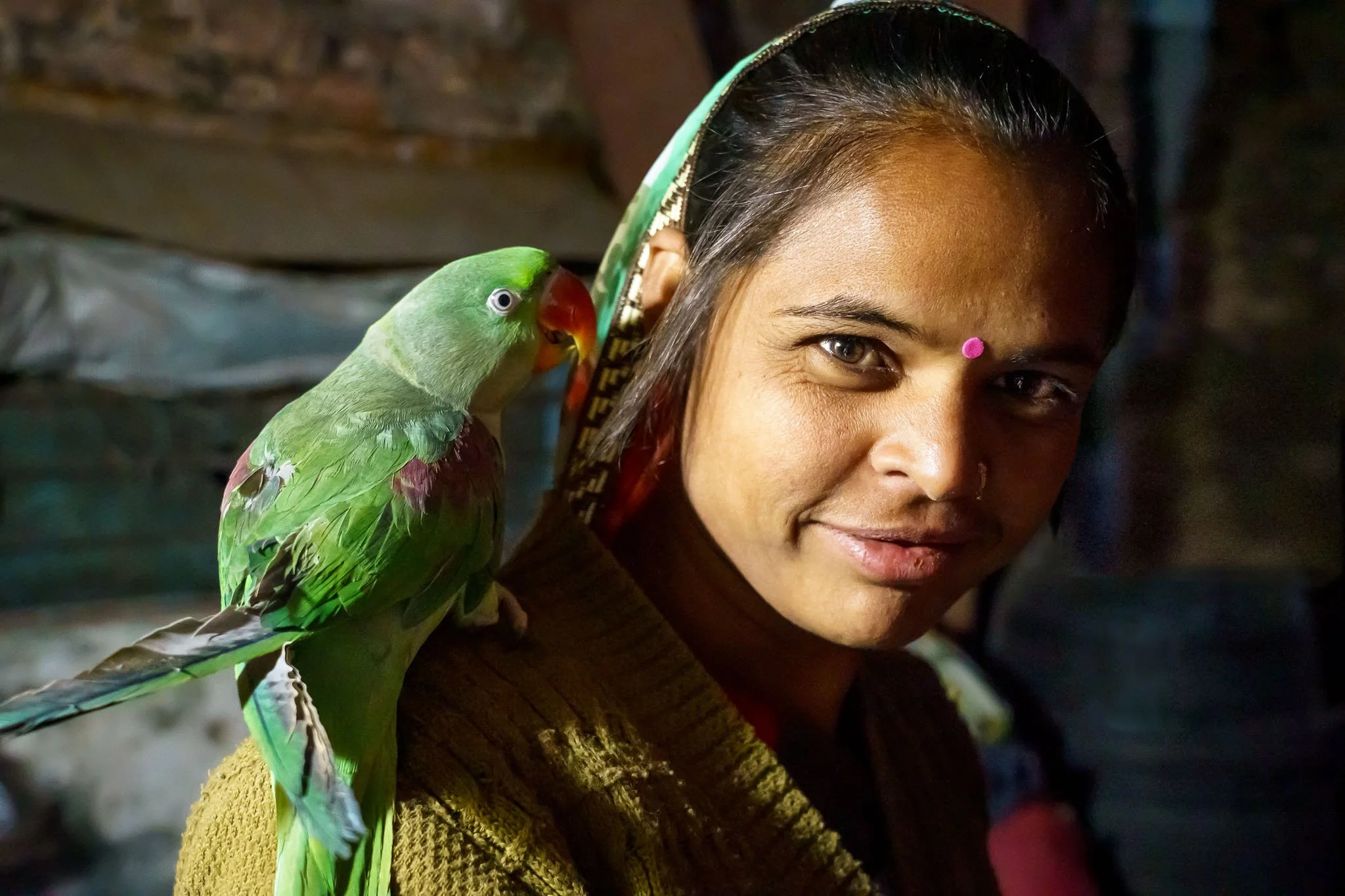 A woman with dark hair, a pink bindi, and a nose ring looks at the camera, with a green parrot perched on her shoulder.