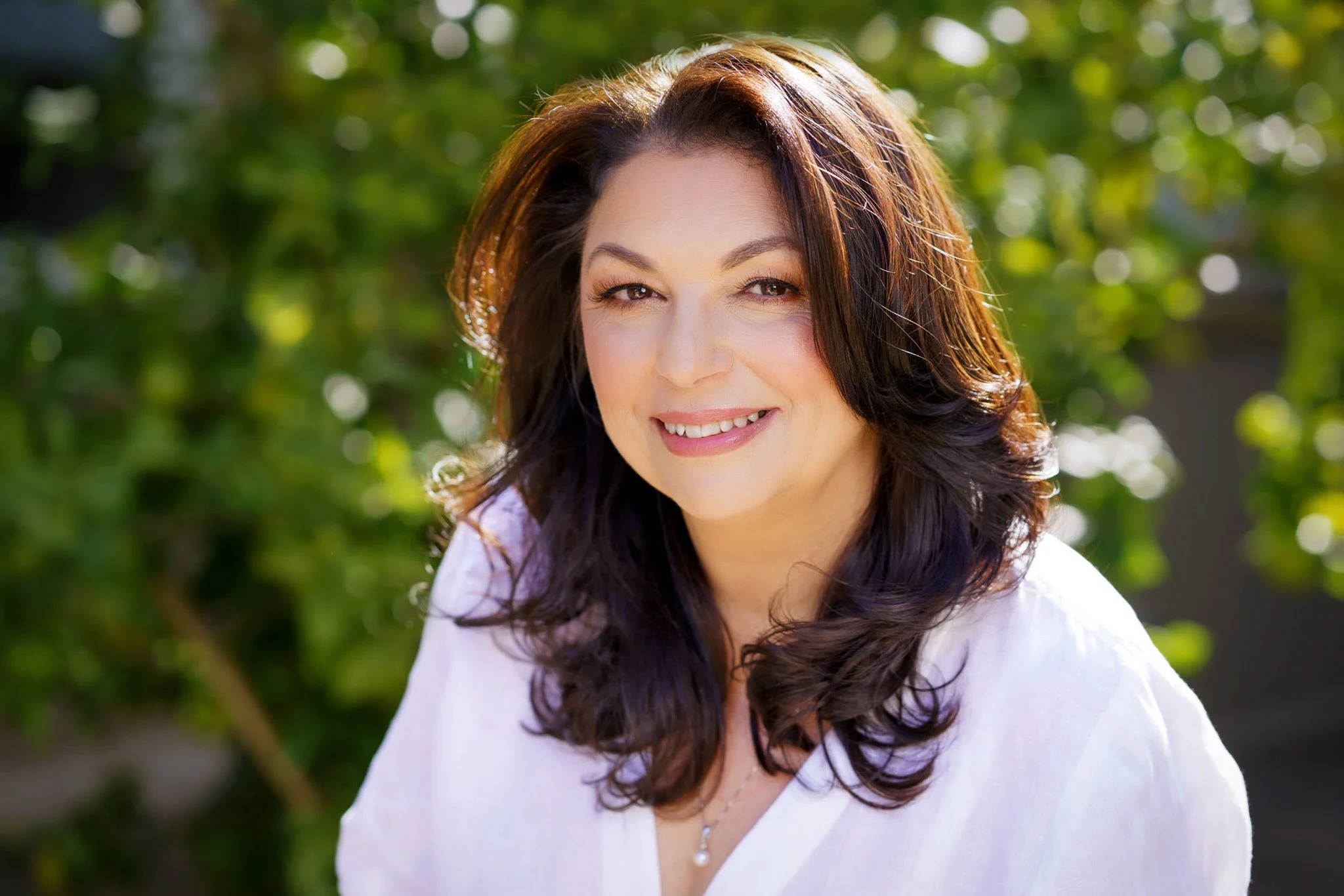 Female headshot taken outdoors against a green backdrop and with the sun backlighting the subject. She is smiling, wearing a white shirt,  and looking relaxed