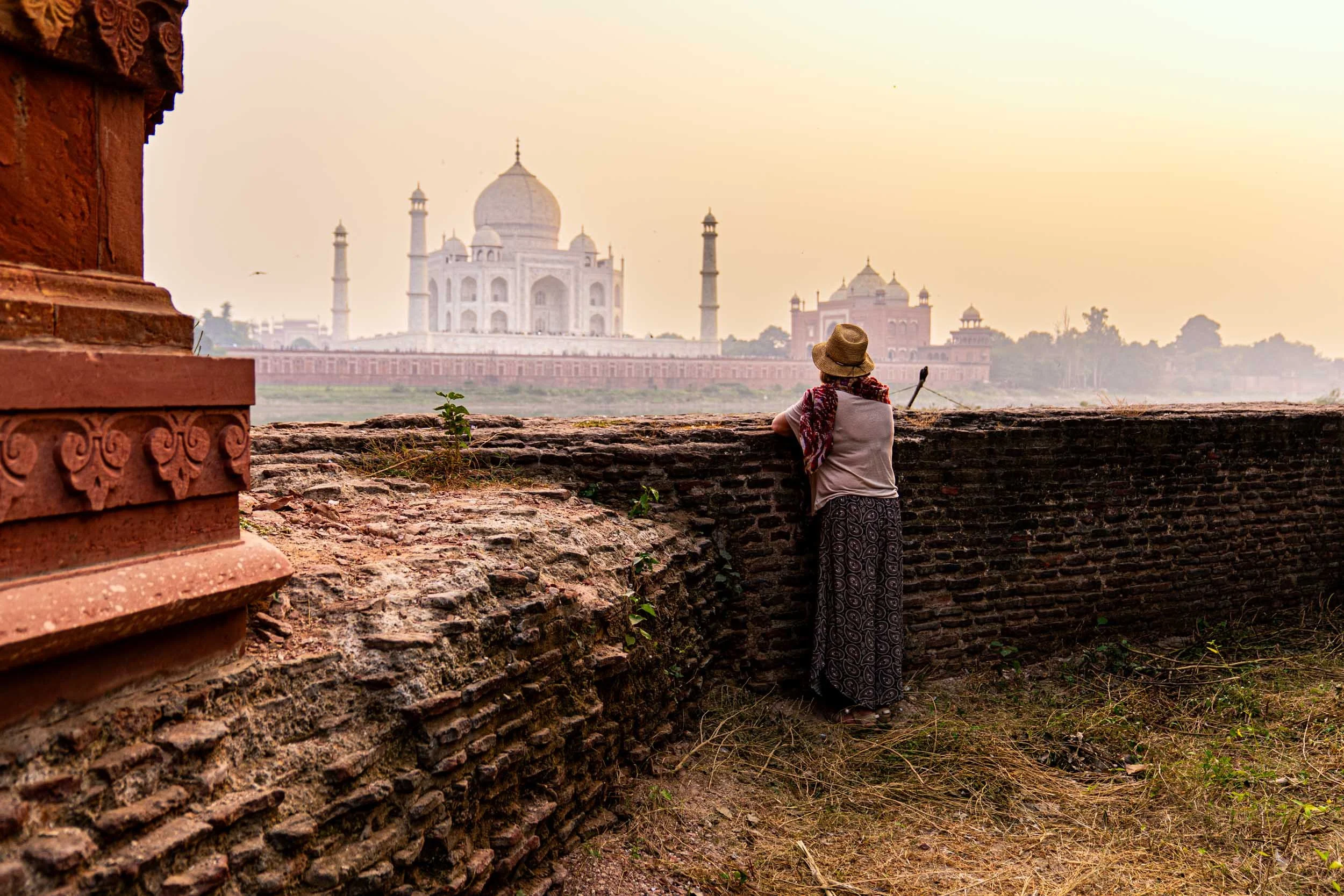 A woman wearing a straw hat, patterned skirt, and a scarf looking at the Taj Mahal through an old brick wall at sunset.