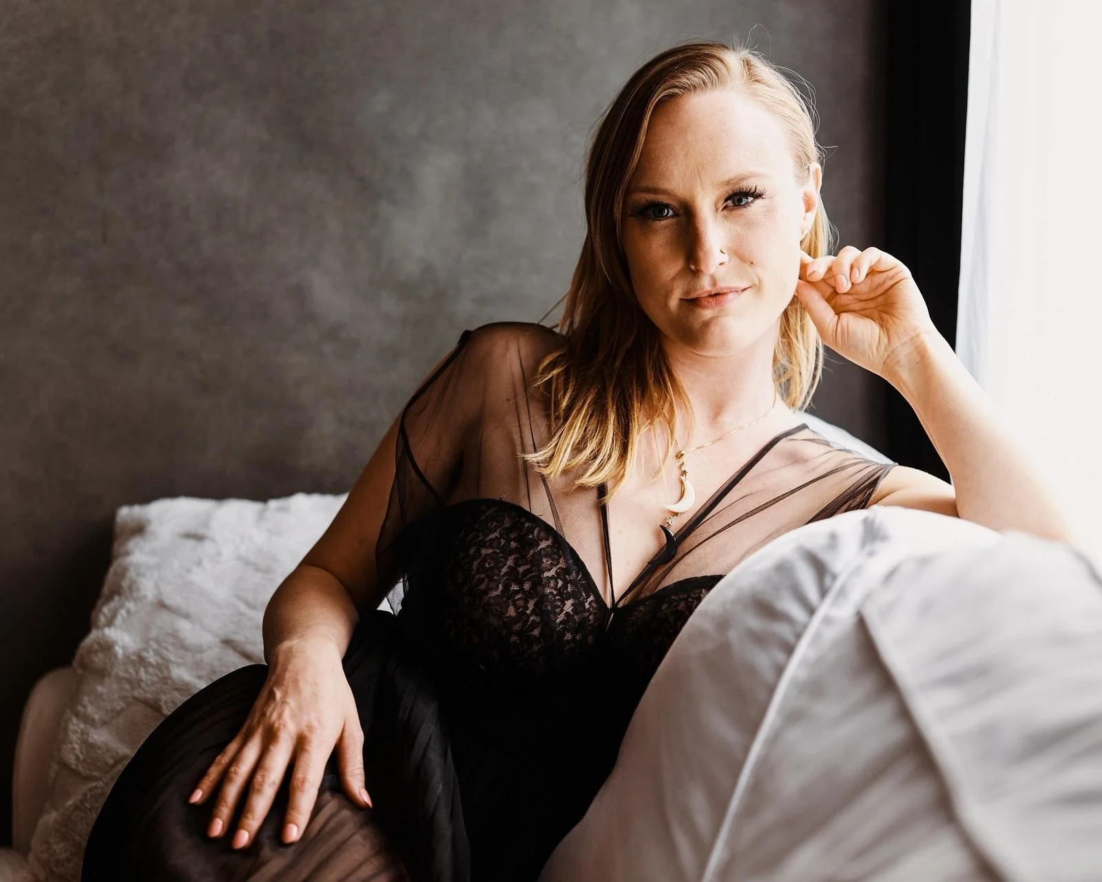 A woman with red hair and a nose piercing sitting on a bed near a window, wearing a sheer black top and black lace lingerie.