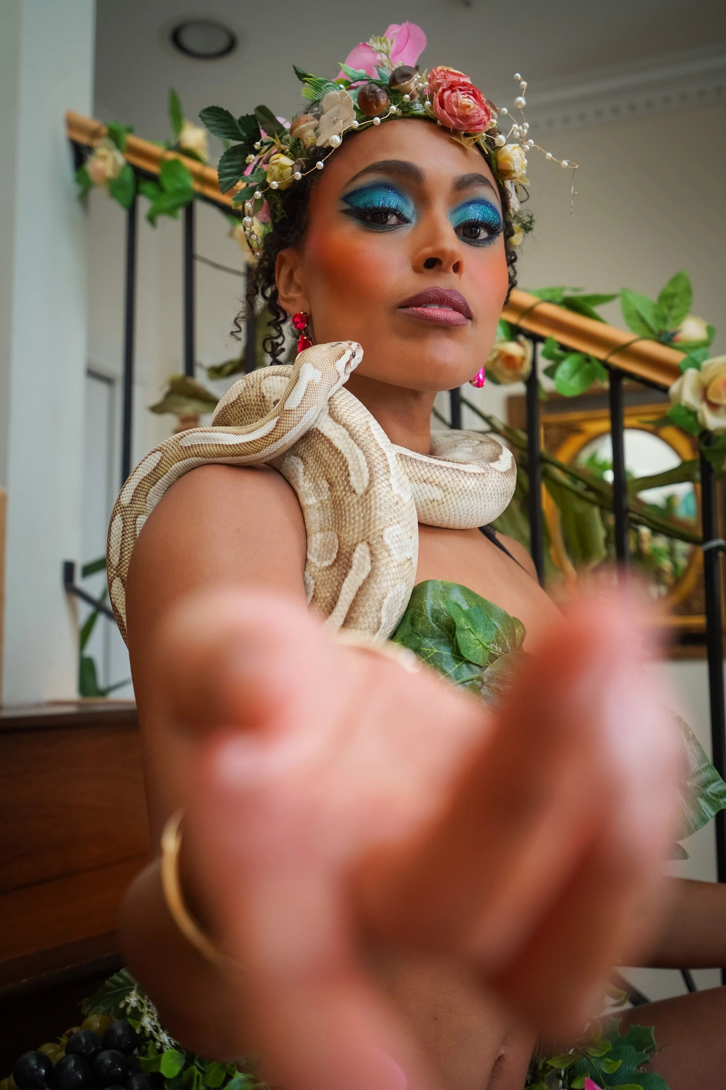A woman with dramatic blue eye makeup, wearing a floral headpiece, pink earrings, and a snake wrapped around her shoulder, standing in front of a floral and black metal background.