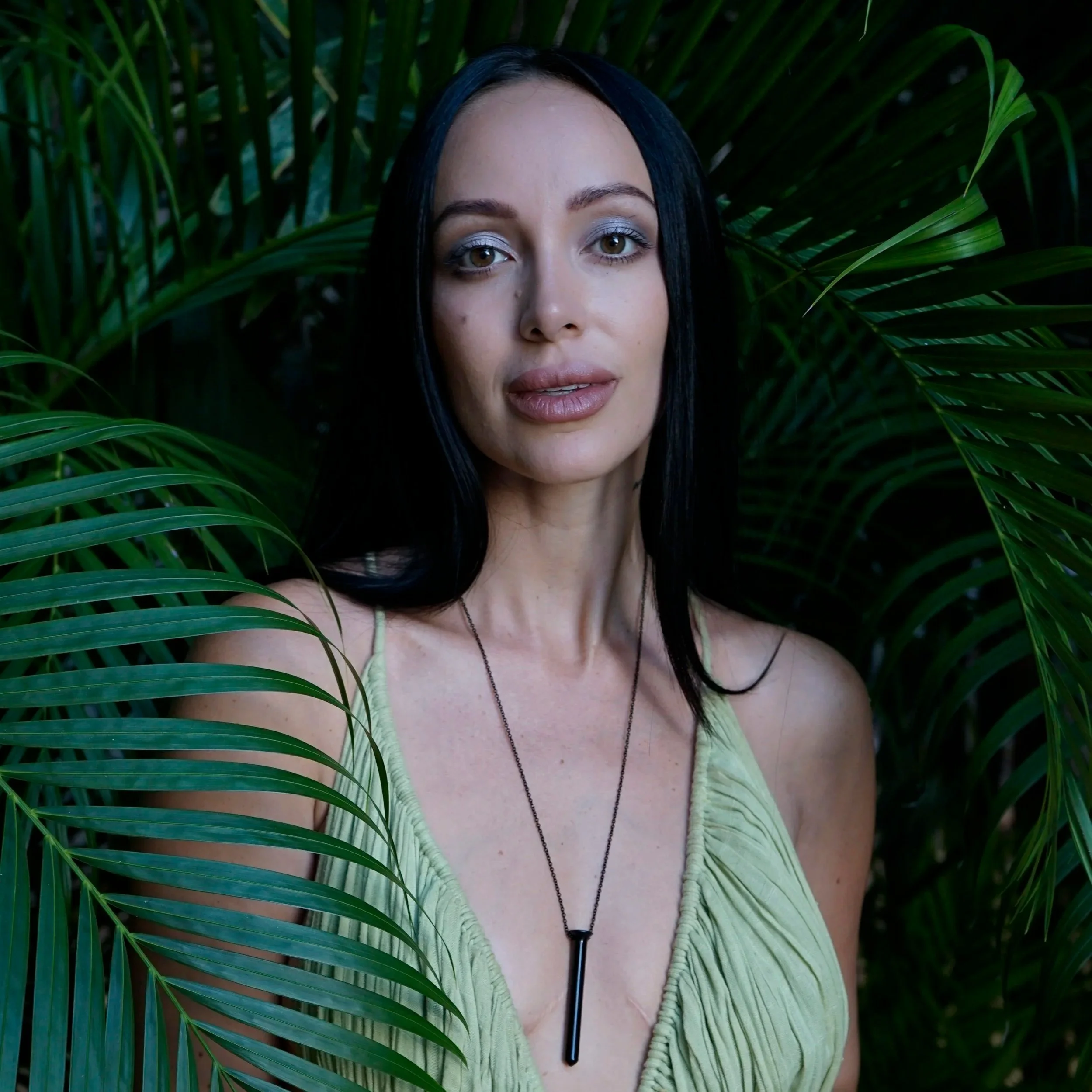 A woman with long dark hair and gray eyes poses among lush green tropical plants, wearing a light green top and a black necklace.