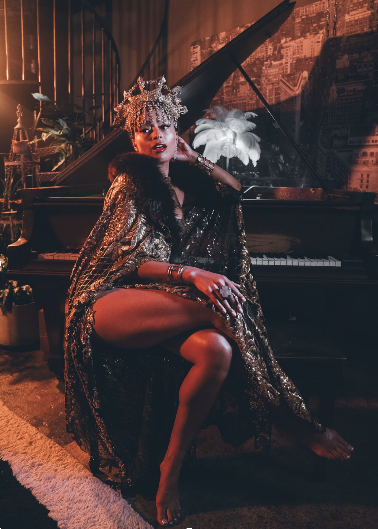 A woman with an ornate crown, gold and black dress, and jewelry sitting on a piano bench, one leg crossed over the other, in a room with plants, a patterned carpet, and a cityscape backdrop.