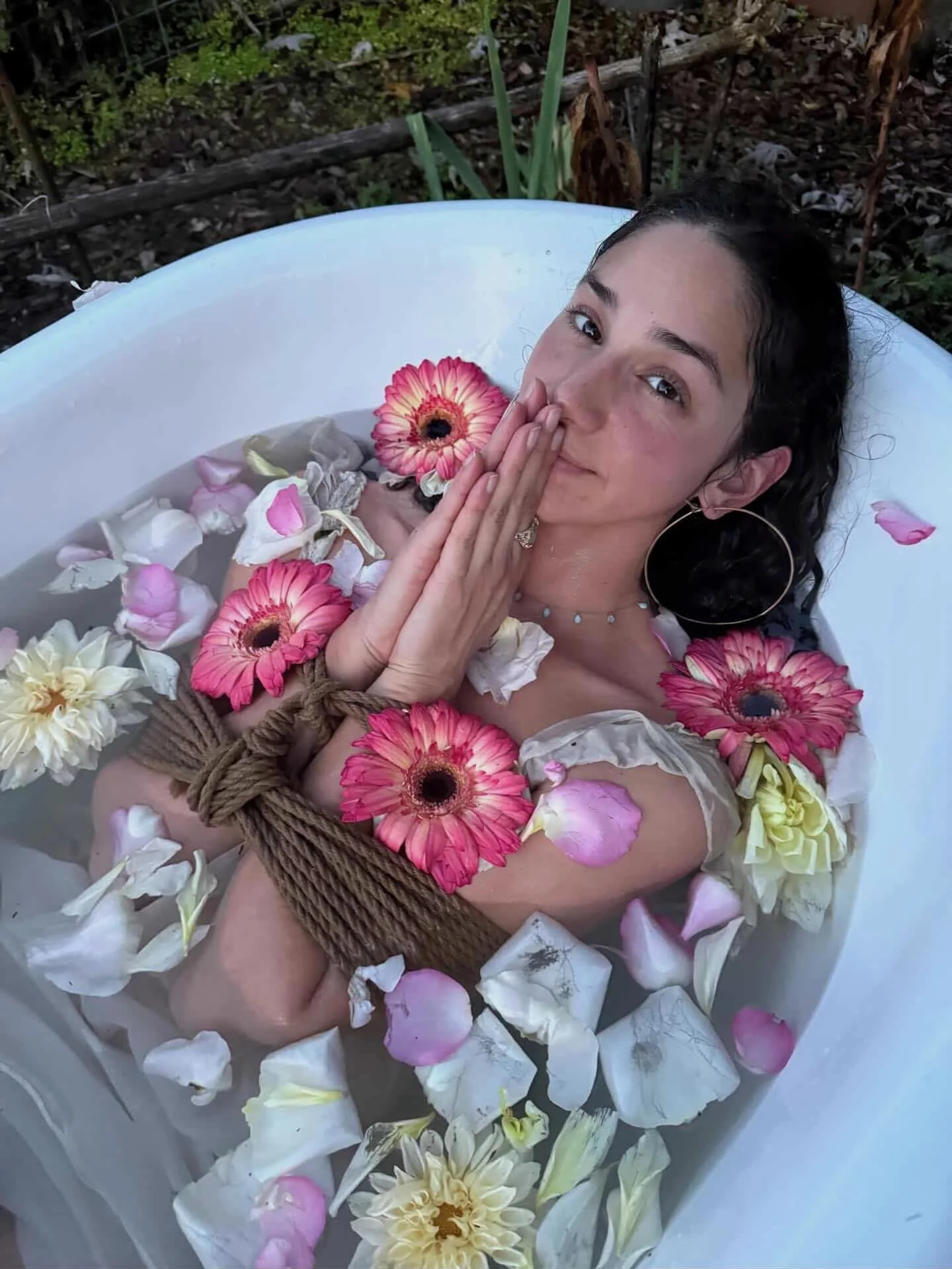 A woman with dark, curly hair in an outdoor bathtub filled with water, surrounded by pink, white, and yellow flowers and rose petals, with green plants and trees in the background.