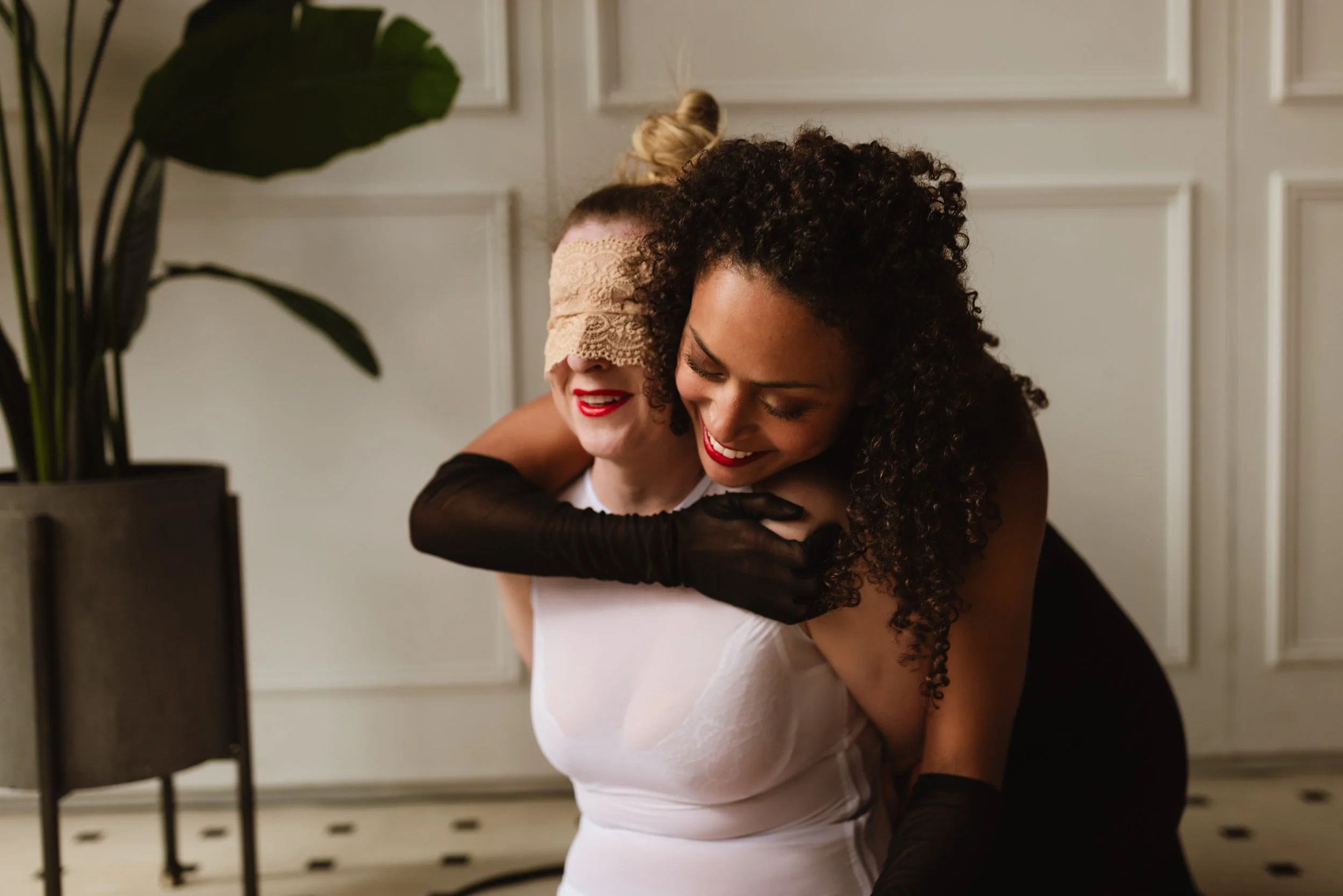 Two women embracing and smiling, one wearing a lace blindfold and the other with curly hair, in a room with white paneled walls and a large green houseplant.