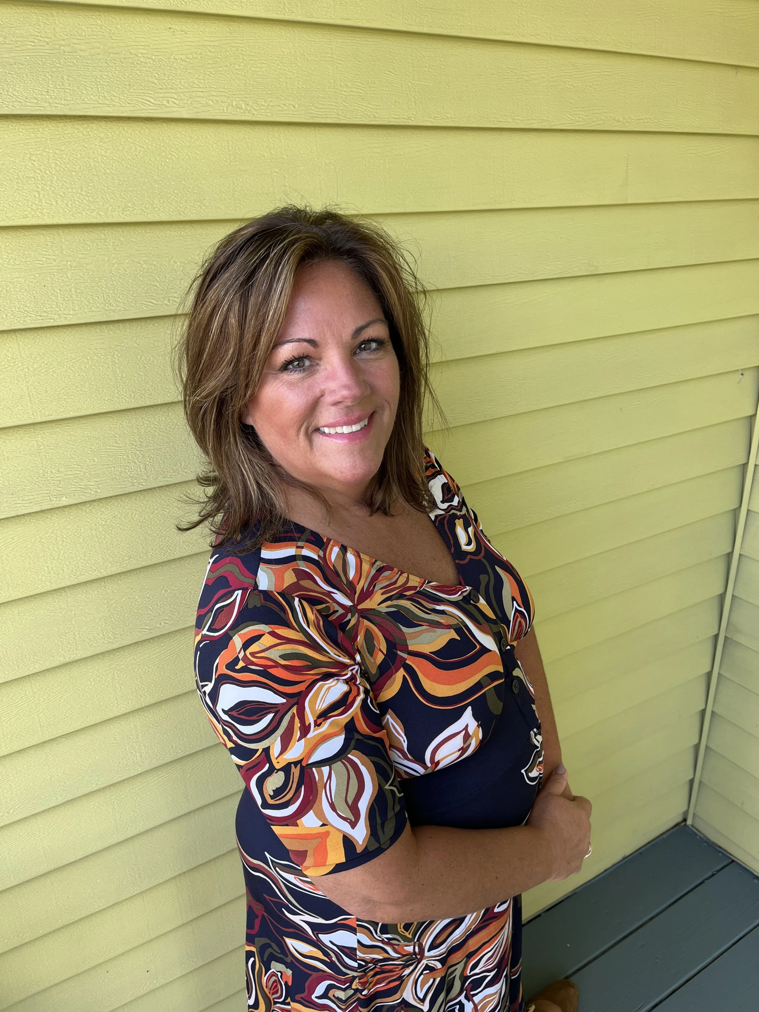 Woman with long brown hair wearing a teal sweater, standing in front of a yellow wall.