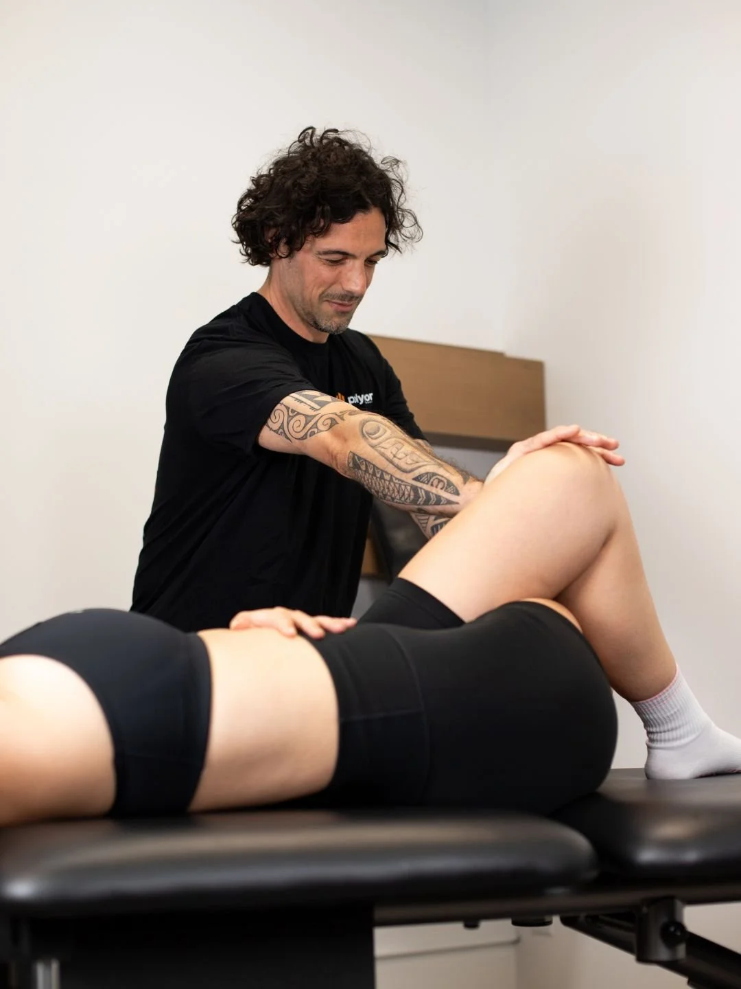 A woman providing physical therapy or injury treatment to a person lying on a table, wearing a black t-shirt with a logo, in a clinical setting.
