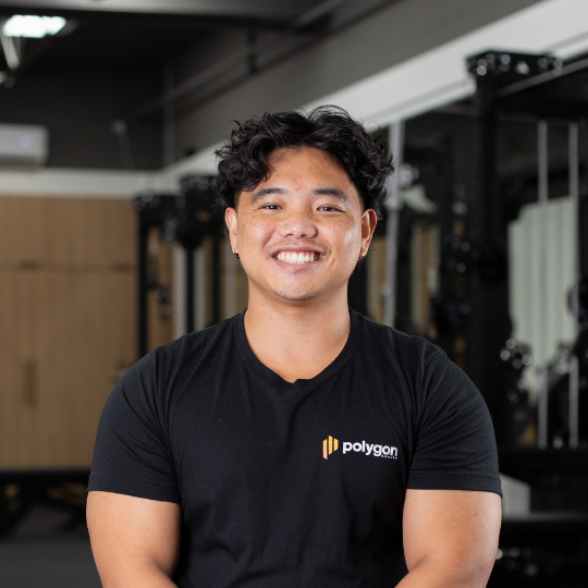 A smiling man with curly black hair and a black t-shirt that says 'polygon' in a gym or fitness studio.
