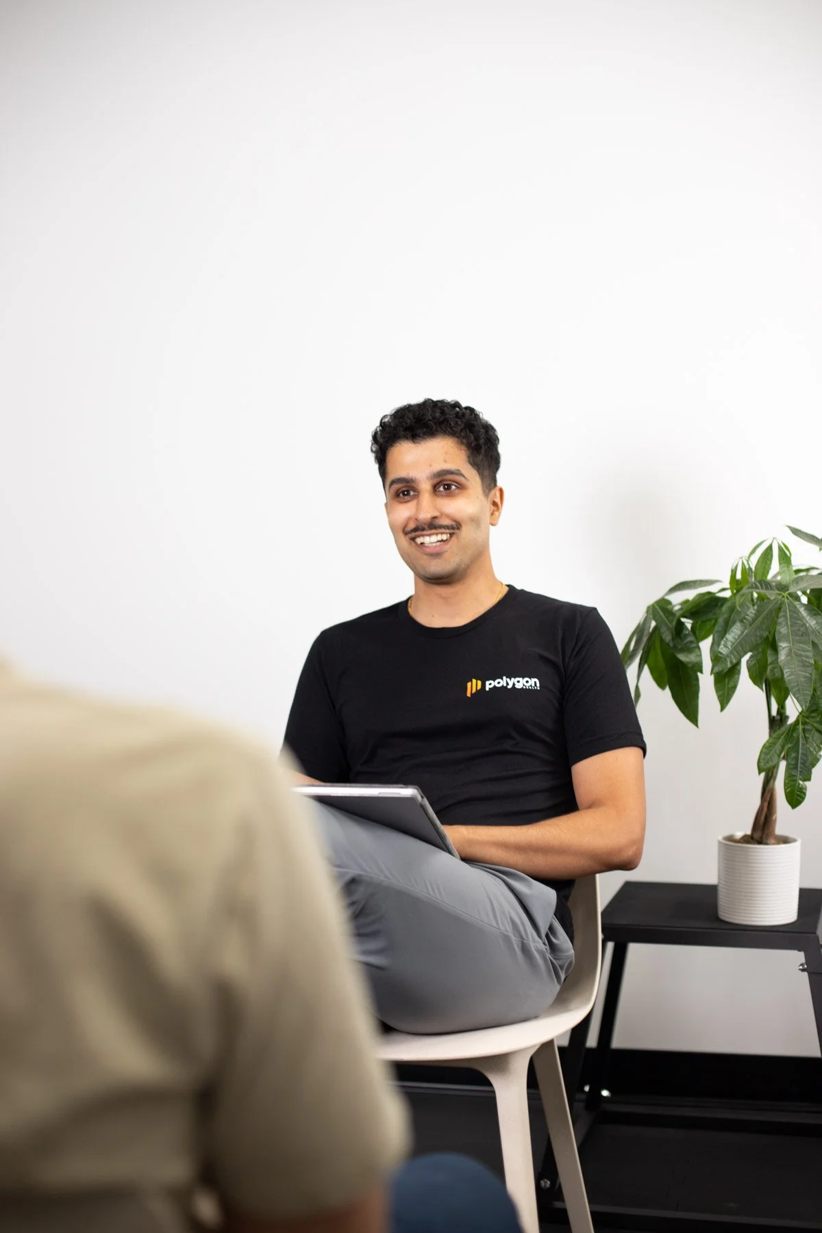 A man sitting in a chair, smiling, holding a notebook, with a woman in the foreground and a potted plant on a table behind him.